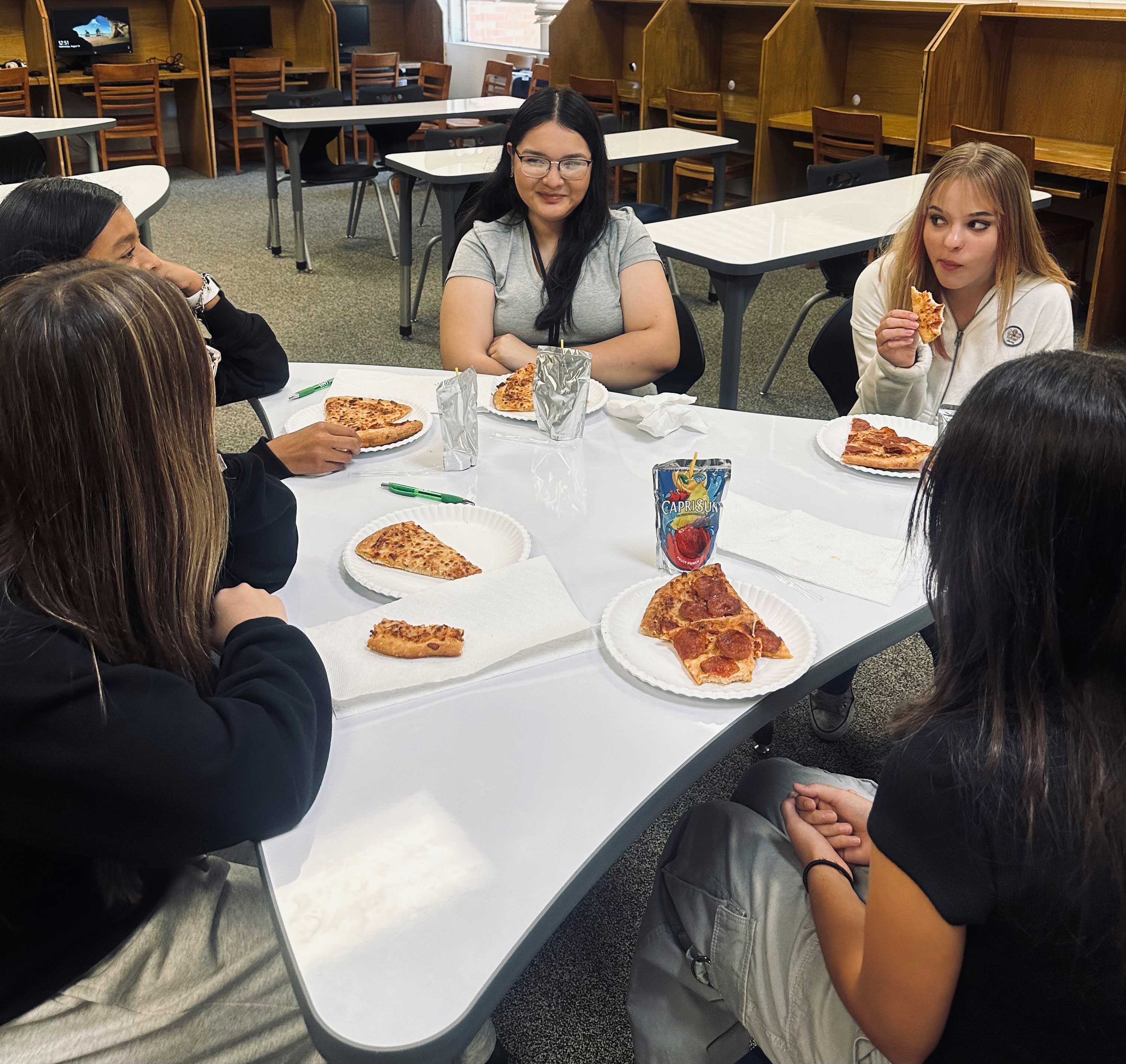 Students sit around a table eating pizza.